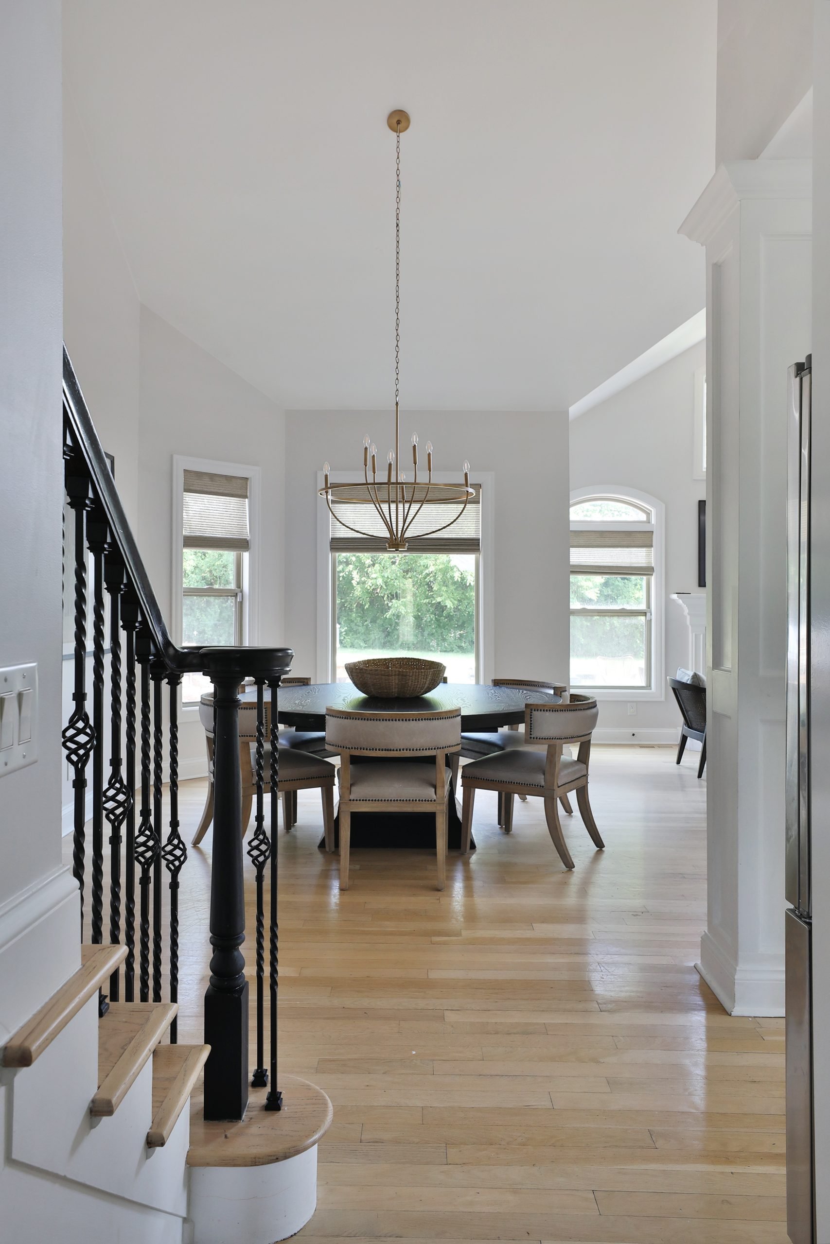 Elegant dining room featuring the Arhaus Leighton Extension Dining Table paired with Carter Dining Chairs from Four Hands. Overhead hangs the Graceful Curve Chandelier in Gold from Shades of Light, adding a warm glow. In the background, tall windows are dressed with soft Roman Shades from Twopages, bringing in natural light and texture to the space.