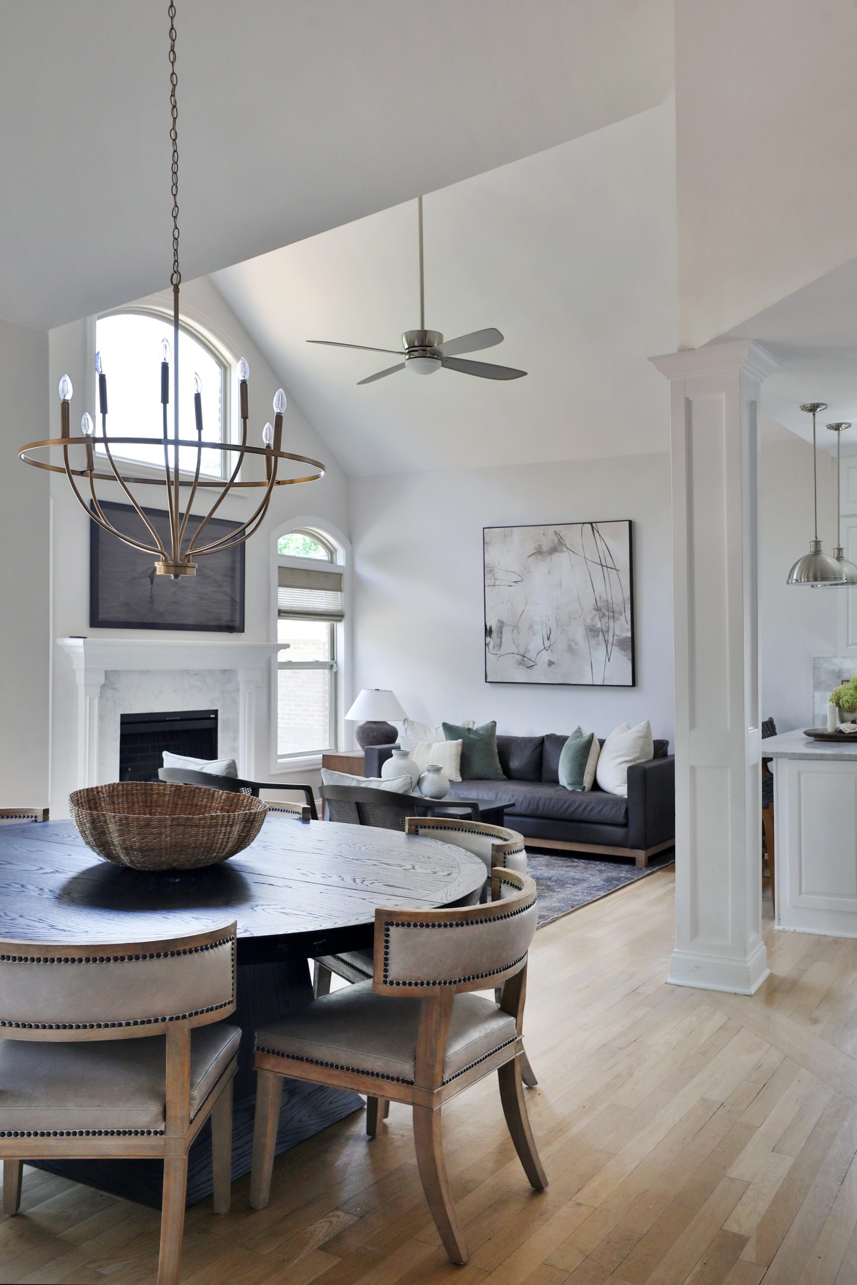 Alternate view of the dining room showcasing the Arhaus Leighton Extension Dining Table and Carter Dining Chairs from Four Hands, centered beneath the Graceful Curve Chandelier in Gold by Shades of Light. In the background, large windows are dressed with Roman Shades from Twopages. A Pottery Barn Jake Leather Sofa sits against the far wall, complemented by a piece of modern artwork hung above, adding depth and contrast to the open-concept space