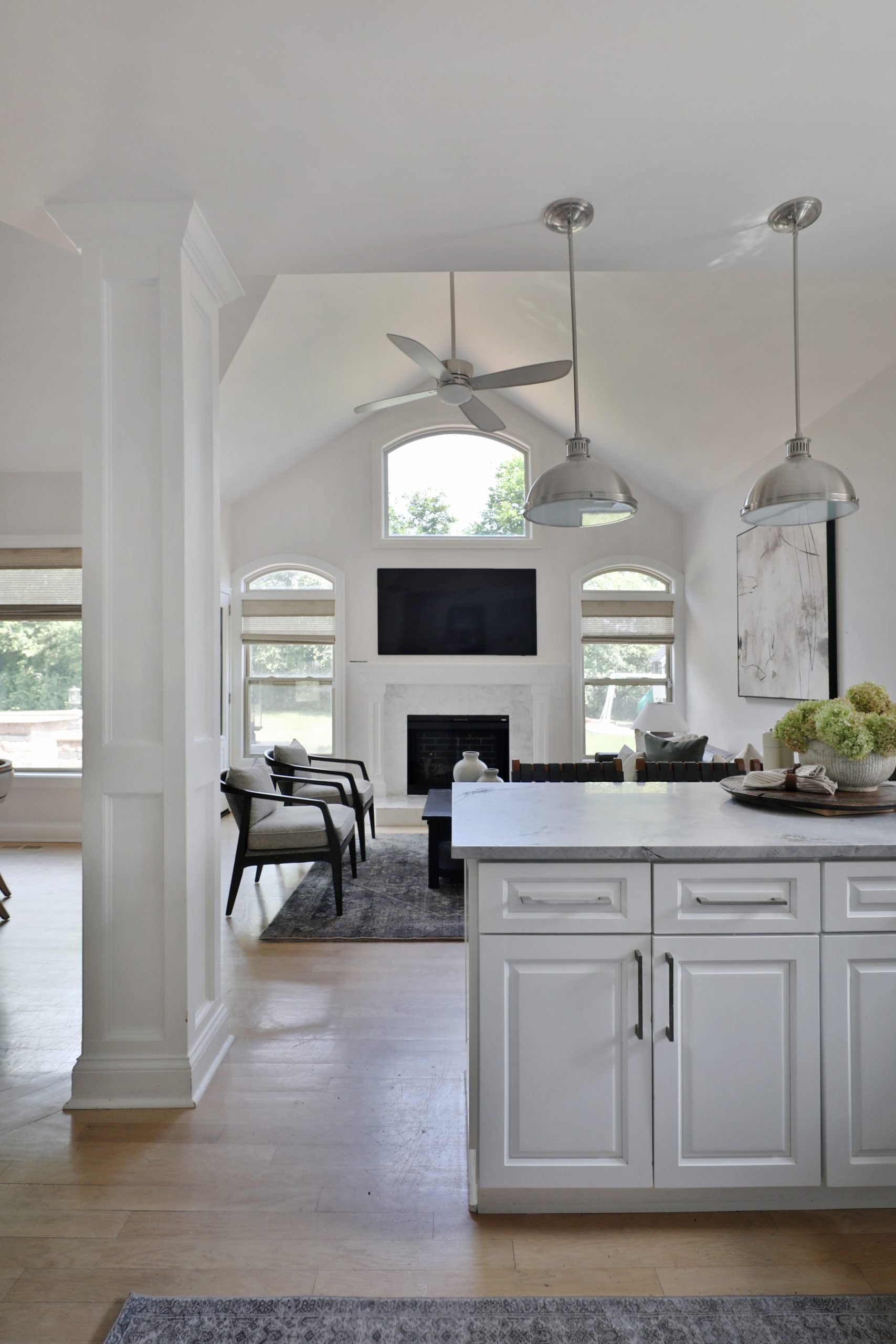 "Bright white kitchen with clean cabinetry and light countertops, opening into a cozy sitting room. The living area features a white fireplace as the focal point, flanked by Roman Shades from Twopages and anchored by an Alexandria Accent Chair from Four Hands