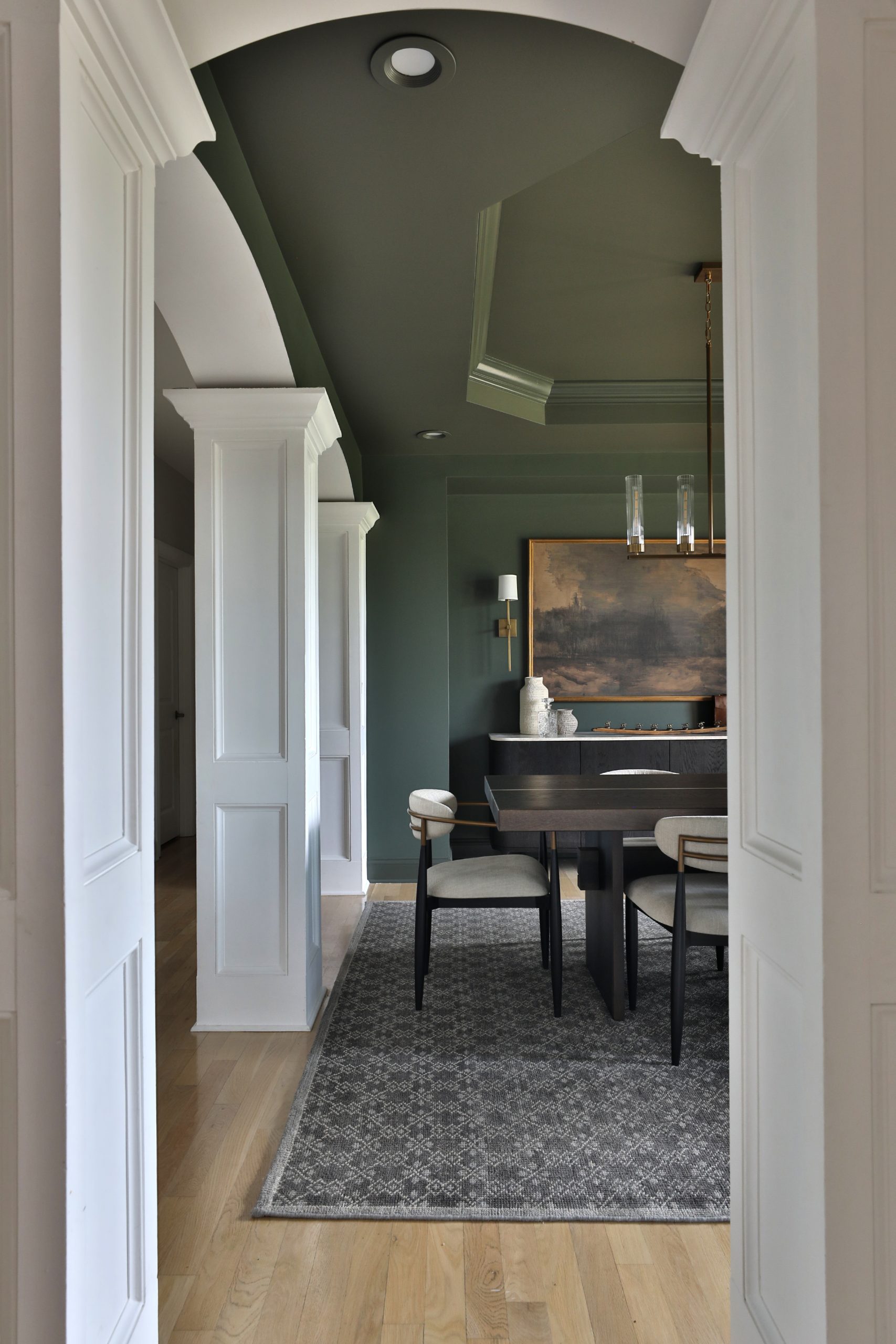 View from foyer into formal dining room with deep green walls, brass chandelier, with a mix of transitional and modern furnishings