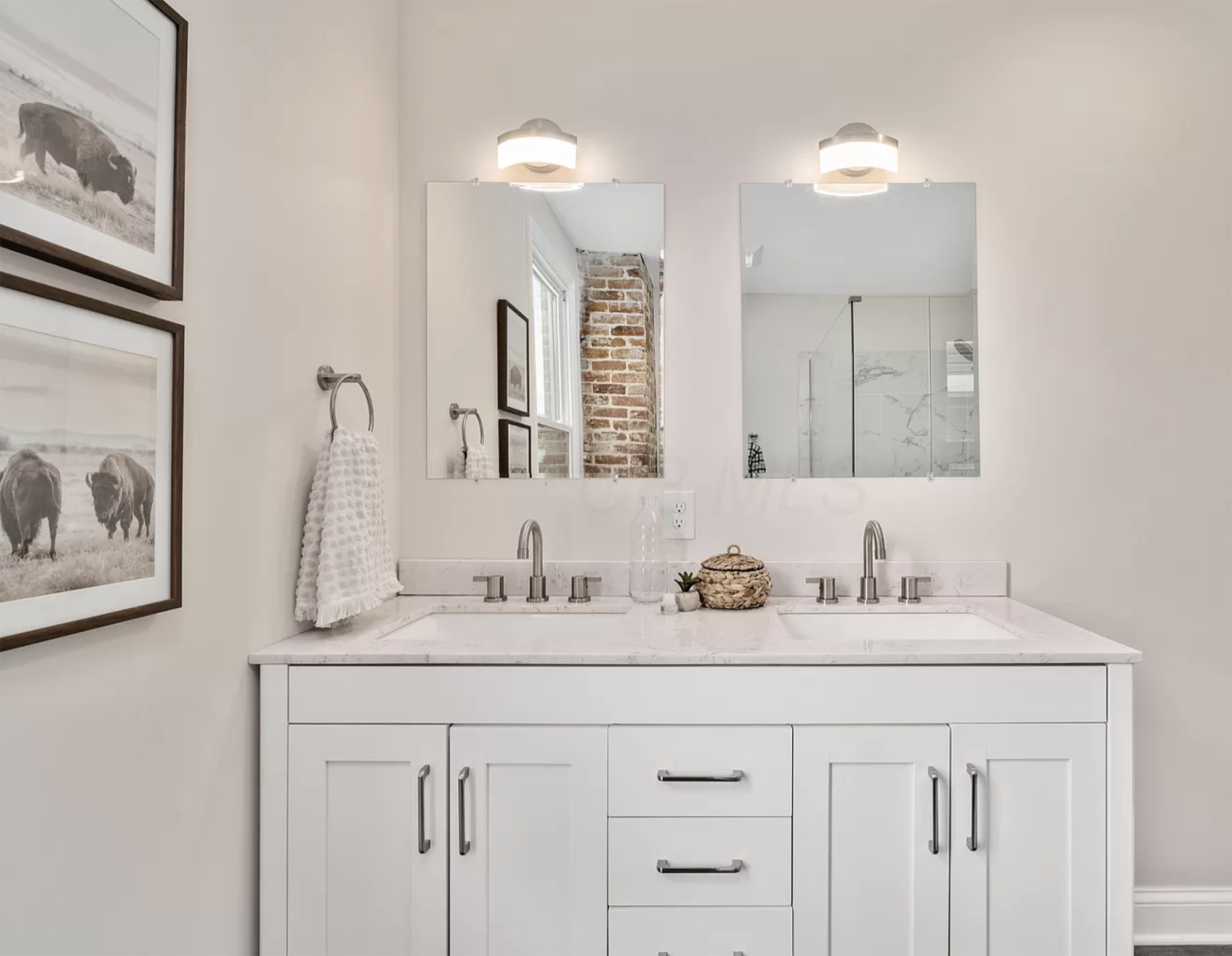 Staged white bathroom design with double vanity, framed wall decor and exposed brick.
